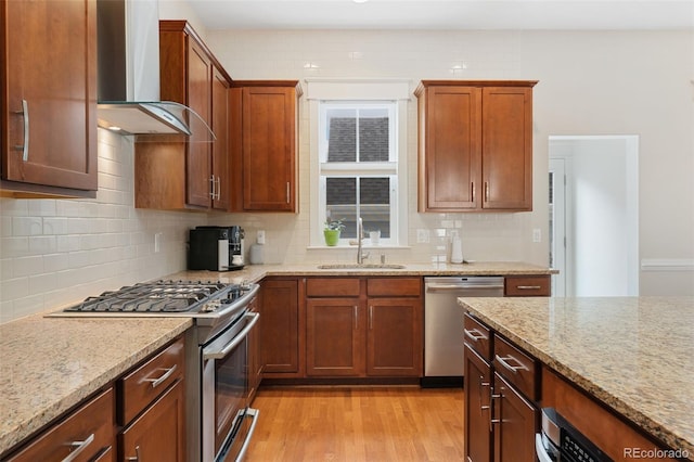 kitchen featuring light stone countertops, light wood-style flooring, a sink, wall chimney exhaust hood, and appliances with stainless steel finishes
