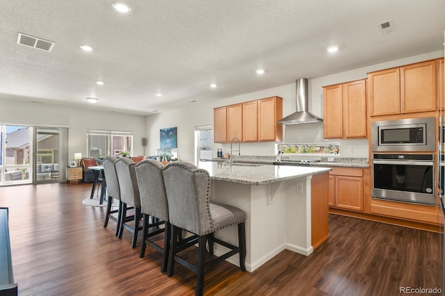 kitchen featuring stainless steel appliances, a sink, dark wood-type flooring, wall chimney exhaust hood, and a kitchen bar