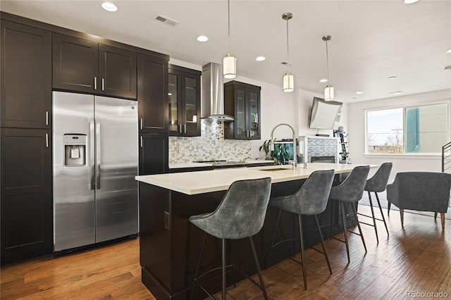 kitchen with light countertops, stainless steel fridge with ice dispenser, a sink, an island with sink, and wall chimney exhaust hood