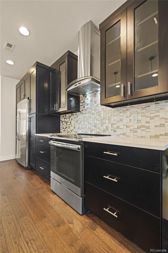 kitchen featuring light countertops, dark wood-style flooring, backsplash, wall chimney range hood, and appliances with stainless steel finishes
