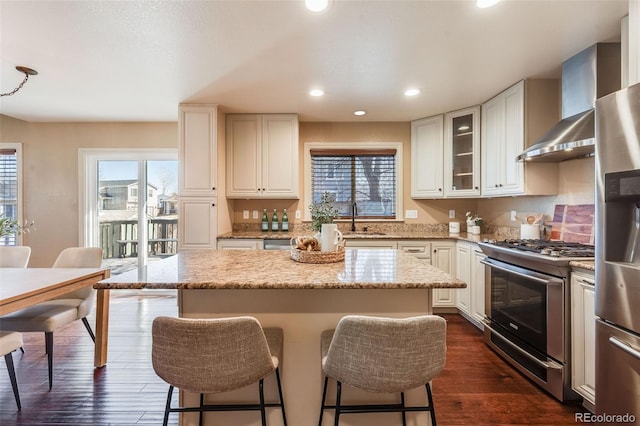 kitchen with a breakfast bar, a sink, stainless steel appliances, dark wood-type flooring, and wall chimney exhaust hood