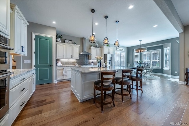 kitchen with wood finished floors, an island with sink, wall chimney range hood, recessed lighting, and tasteful backsplash