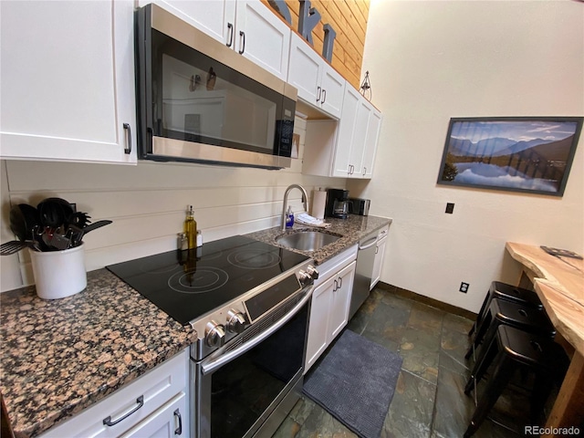 kitchen featuring stone tile floors, stainless steel appliances, white cabinetry, a sink, and dark stone counters