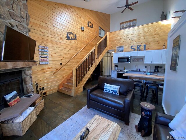 living area featuring a fireplace, a towering ceiling, ceiling fan, wood walls, and stairs