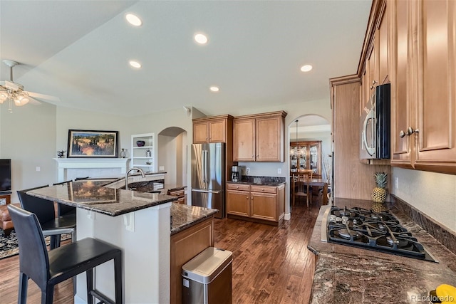 kitchen with appliances with stainless steel finishes, ceiling fan, a center island with sink, dark hardwood / wood-style floors, and dark stone countertops