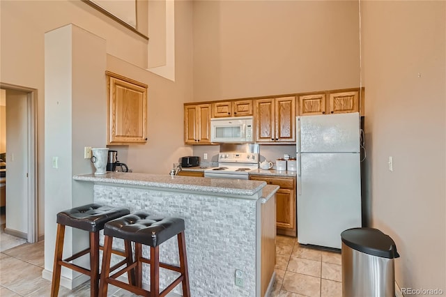 kitchen featuring a kitchen bar, kitchen peninsula, a towering ceiling, white appliances, and light tile patterned flooring