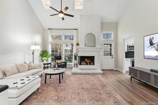 living room featuring a ceiling fan, a skylight, a brick fireplace, and high vaulted ceiling