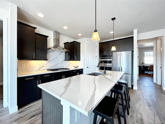 kitchen with appliances with stainless steel finishes, pendant lighting, an island with sink, sink, and wall chimney range hood