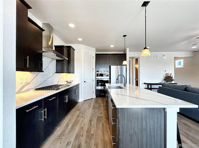 kitchen with appliances with stainless steel finishes, pendant lighting, sink, a large island with sink, and wall chimney range hood