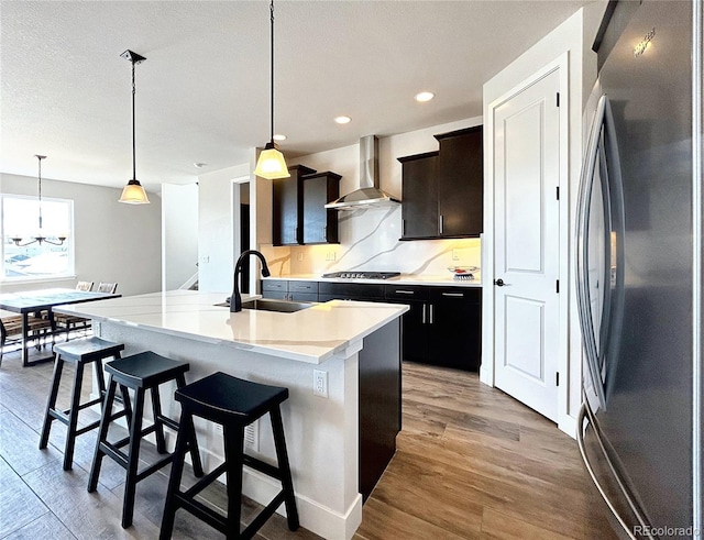 kitchen featuring wall chimney exhaust hood, sink, appliances with stainless steel finishes, an island with sink, and pendant lighting