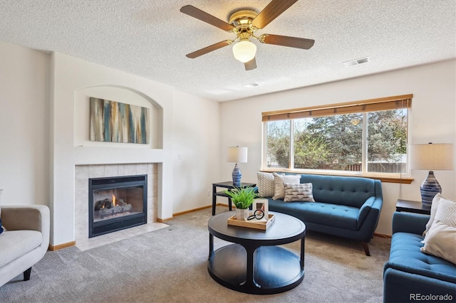 carpeted living room featuring ceiling fan, a textured ceiling, a tiled fireplace, baseboards, and visible vents
