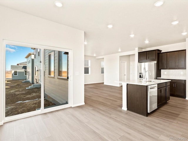 kitchen featuring tasteful backsplash, light wood-type flooring, appliances with stainless steel finishes, an island with sink, and dark brown cabinets