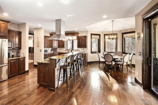 kitchen with a kitchen bar, dark wood-style floors, freestanding refrigerator, island range hood, and light stone countertops