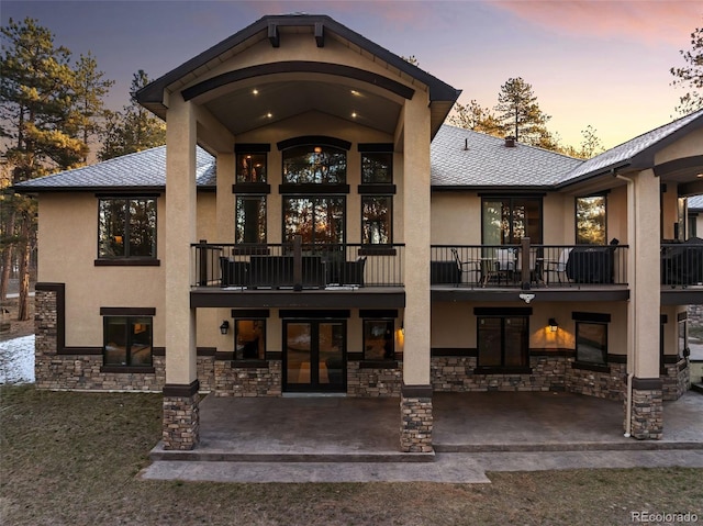 back of house at dusk featuring a balcony, stone siding, stucco siding, and a patio