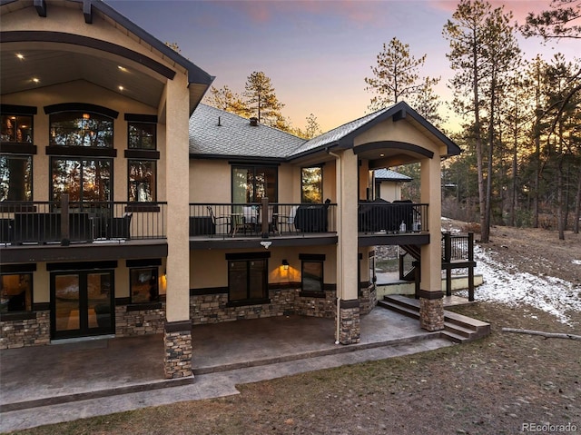 rear view of property with stucco siding, stone siding, a shingled roof, stairs, and a patio area