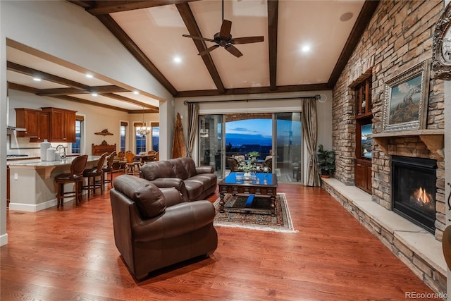 living room featuring high vaulted ceiling, light wood-style flooring, a stone fireplace, beamed ceiling, and ceiling fan with notable chandelier