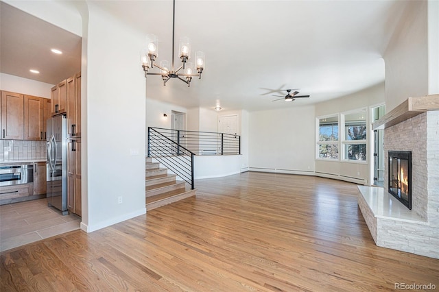 unfurnished living room featuring a baseboard radiator, light wood-style flooring, ceiling fan with notable chandelier, stairs, and a glass covered fireplace