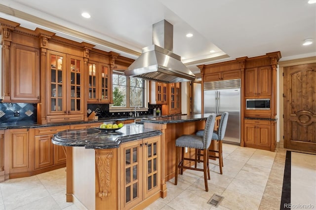 kitchen with island range hood, a kitchen island, glass insert cabinets, a breakfast bar area, and built in appliances