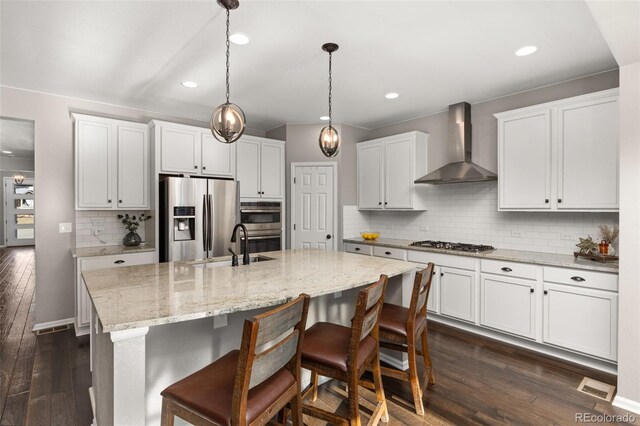 kitchen with a kitchen island with sink, white cabinets, wall chimney range hood, appliances with stainless steel finishes, and tasteful backsplash