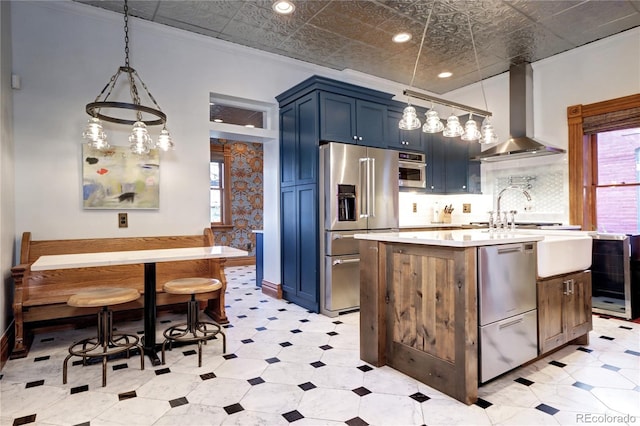 kitchen featuring blue cabinetry, wall chimney range hood, decorative light fixtures, a center island with sink, and appliances with stainless steel finishes