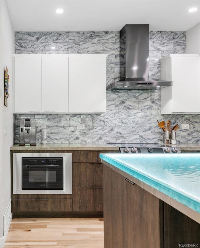 kitchen with wall chimney range hood, white cabinets, light wood-style floors, and tasteful backsplash