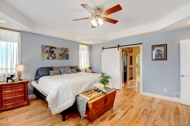 bedroom featuring a barn door, ceiling fan, a raised ceiling, and light wood-type flooring