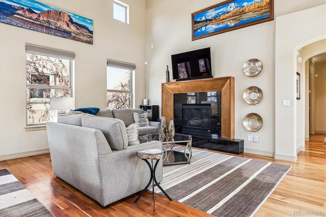 living room featuring arched walkways, wood finished floors, a towering ceiling, baseboards, and a tiled fireplace