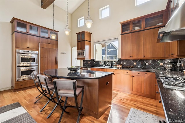 kitchen with light wood finished floors, glass insert cabinets, appliances with stainless steel finishes, a kitchen island, and ventilation hood