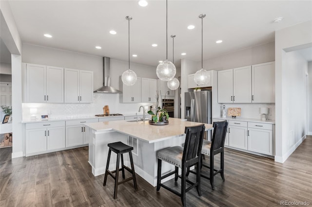 kitchen with stainless steel refrigerator with ice dispenser, light countertops, a kitchen island with sink, white cabinets, and wall chimney range hood