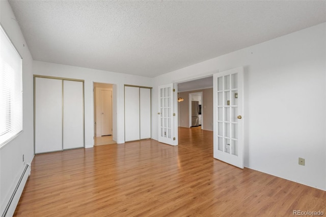 unfurnished bedroom featuring french doors, baseboard heating, multiple closets, light wood-style flooring, and a textured ceiling