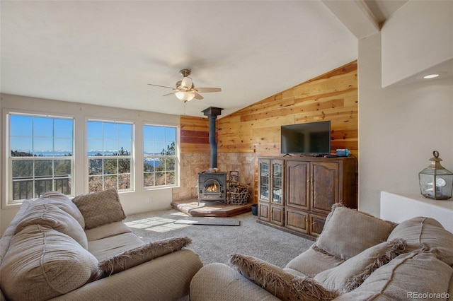 living room with wooden walls, lofted ceiling, ceiling fan, a wood stove, and carpet flooring