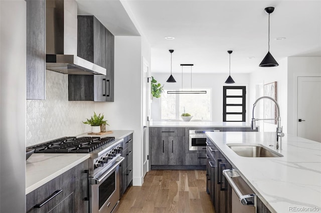 kitchen with appliances with stainless steel finishes, sink, wall chimney range hood, light hardwood / wood-style flooring, and hanging light fixtures