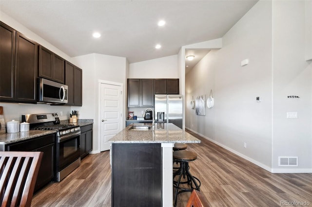 kitchen with dark brown cabinetry, sink, stainless steel appliances, light stone countertops, and a kitchen island with sink