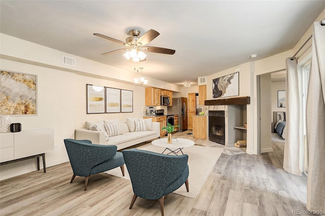 living room featuring light wood-style floors, a fireplace, visible vents, and ceiling fan with notable chandelier
