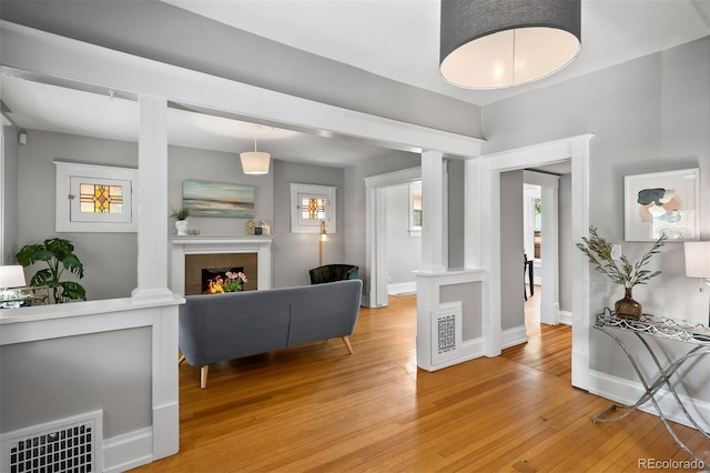 living area featuring light wood-style floors, baseboards, a warm lit fireplace, and ornate columns