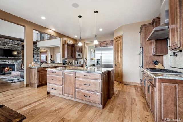 kitchen with pendant lighting, built in appliances, a fireplace, an island with sink, and wall chimney exhaust hood