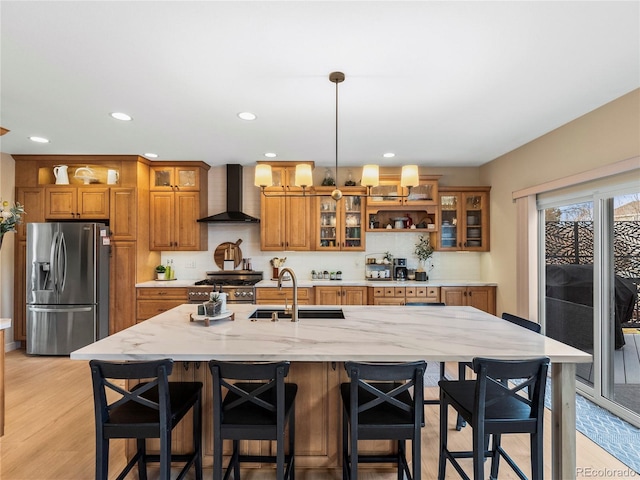 kitchen featuring light wood finished floors, wall chimney exhaust hood, appliances with stainless steel finishes, brown cabinets, and a sink