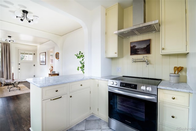 kitchen featuring wall chimney range hood, stainless steel range with electric cooktop, light stone counters, and kitchen peninsula