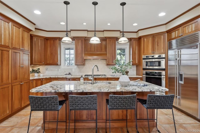 kitchen featuring brown cabinets, stainless steel appliances, backsplash, a sink, and an island with sink