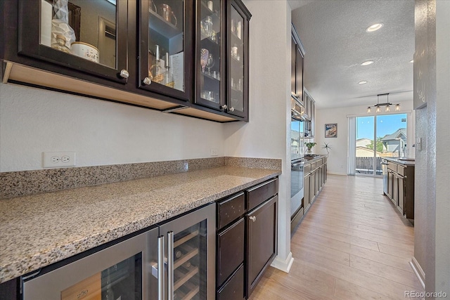 bar with light stone countertops, a textured ceiling, dark brown cabinetry, wine cooler, and light hardwood / wood-style flooring