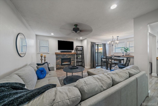 living area featuring light wood finished floors, ceiling fan with notable chandelier, a textured ceiling, baseboards, and a stone fireplace