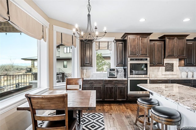 kitchen featuring dark brown cabinetry, hanging light fixtures, light stone counters, and double oven