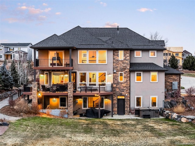 back house at dusk featuring a hot tub, a patio, a balcony, and a yard