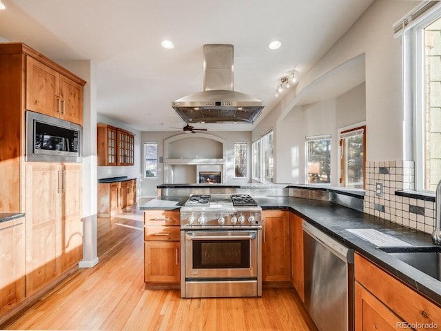 kitchen with island exhaust hood, light wood-type flooring, ceiling fan, stainless steel appliances, and decorative backsplash