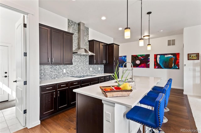 kitchen featuring hardwood / wood-style floors, black electric stovetop, wall chimney exhaust hood, decorative light fixtures, and a breakfast bar area