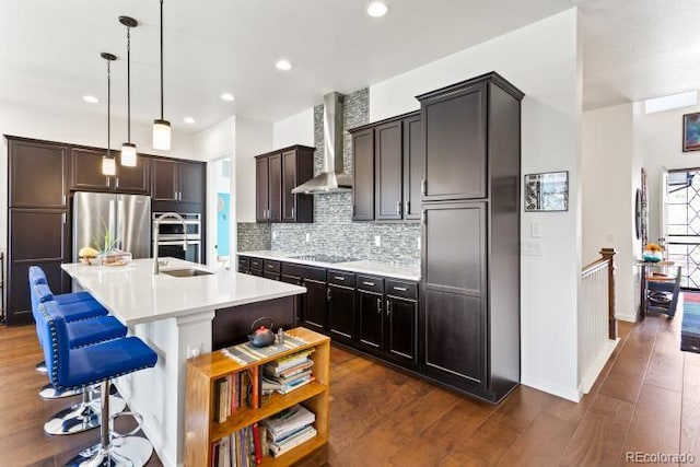 kitchen featuring wall chimney range hood, dark hardwood / wood-style floors, pendant lighting, a kitchen island with sink, and appliances with stainless steel finishes