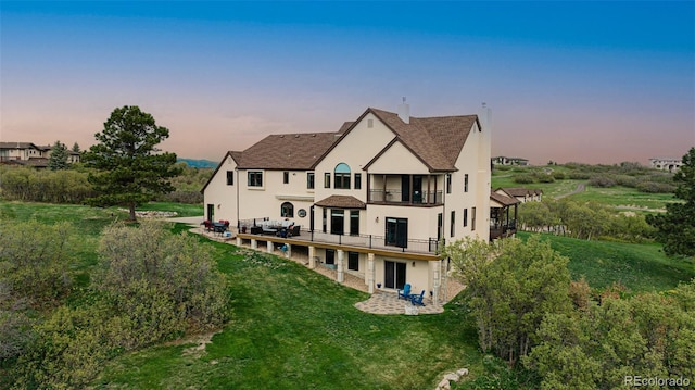 back of property at dusk featuring a balcony, stucco siding, a yard, a patio area, and a shingled roof