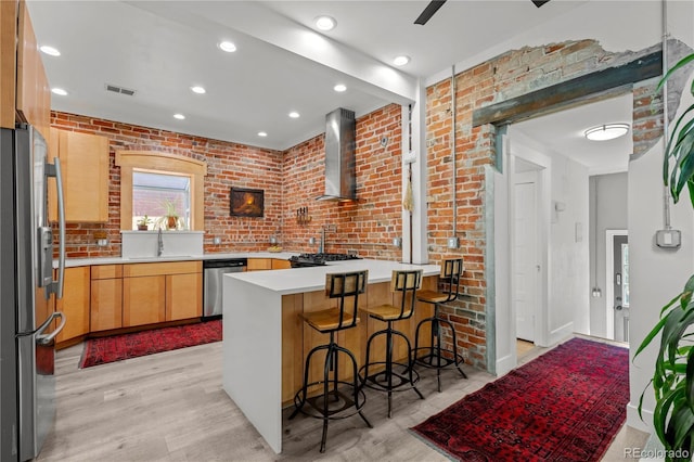 kitchen featuring stainless steel appliances, wall chimney exhaust hood, a peninsula, light wood-style floors, and a sink