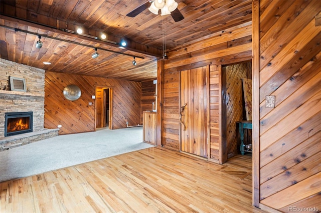 unfurnished living room with a stone fireplace, wood walls, a ceiling fan, wood ceiling, and light wood-style floors