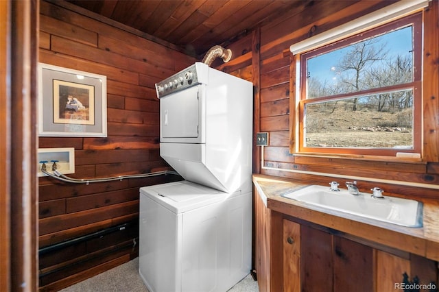 clothes washing area featuring laundry area, wood walls, stacked washing maching and dryer, and a sink